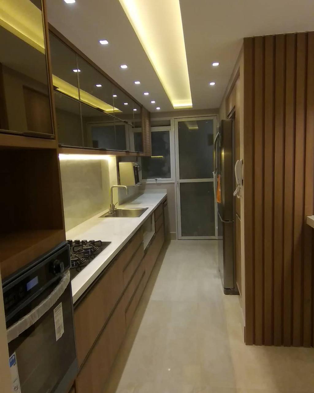 Cozinha residencial com acabamento premium: marcenaria sob medida em madeira, bancadas em pedra, iluminação embutida em sanca e integração de eletrodomésticos.