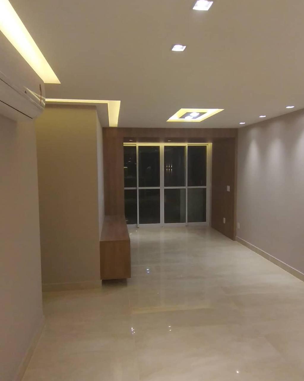 Interior residencial de alto padrão com piso porcelanato polido, forro de gesso com iluminação embutida, painel em madeira nobre e esquadrias de vidro.