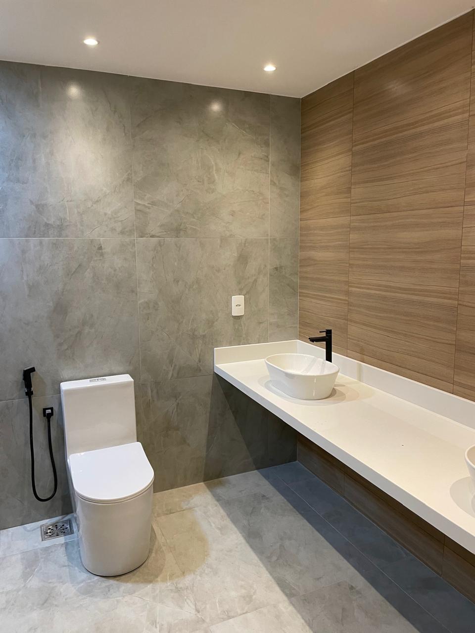 Banheiro com acabamento premium, revestimento em porcelanato cinza mármore, parede em porcelanato amadeirado, louças brancas e metais pretos.