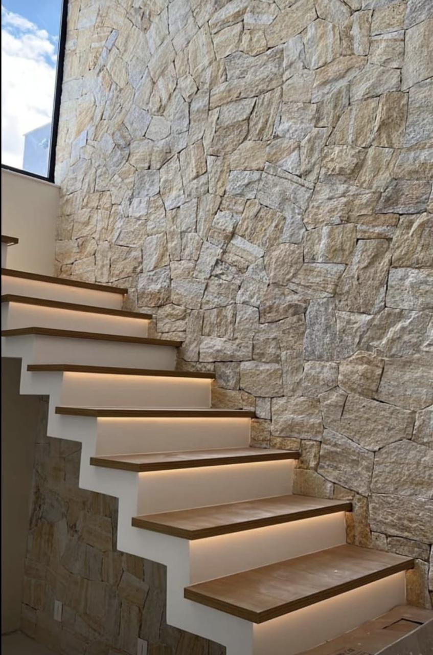 Escada interna de alto padrão com parede em pedra natural, degraus em madeira e iluminação LED embutida sob os espelhos, acabamento premium impecável.