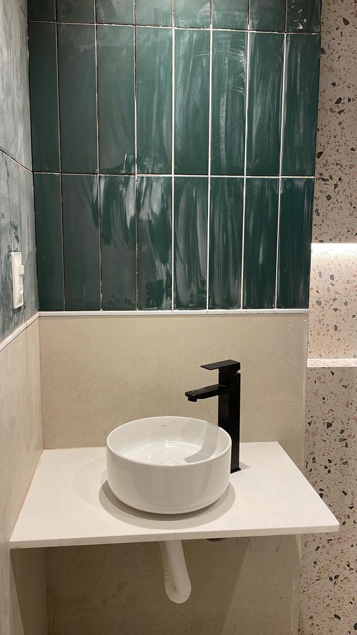Banheiro com acabamento premium: parede de azulejos verdes brilhantes, bancada com cuba esmaltada, torneira preta e parede em terrazzo.