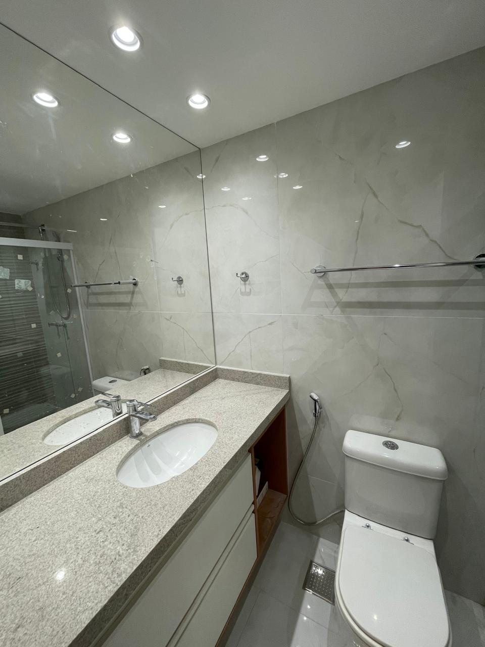 Banheiro de alto padrão com bancada em granito, gabinete branco, espelho amplo e revestimento marmorizado.