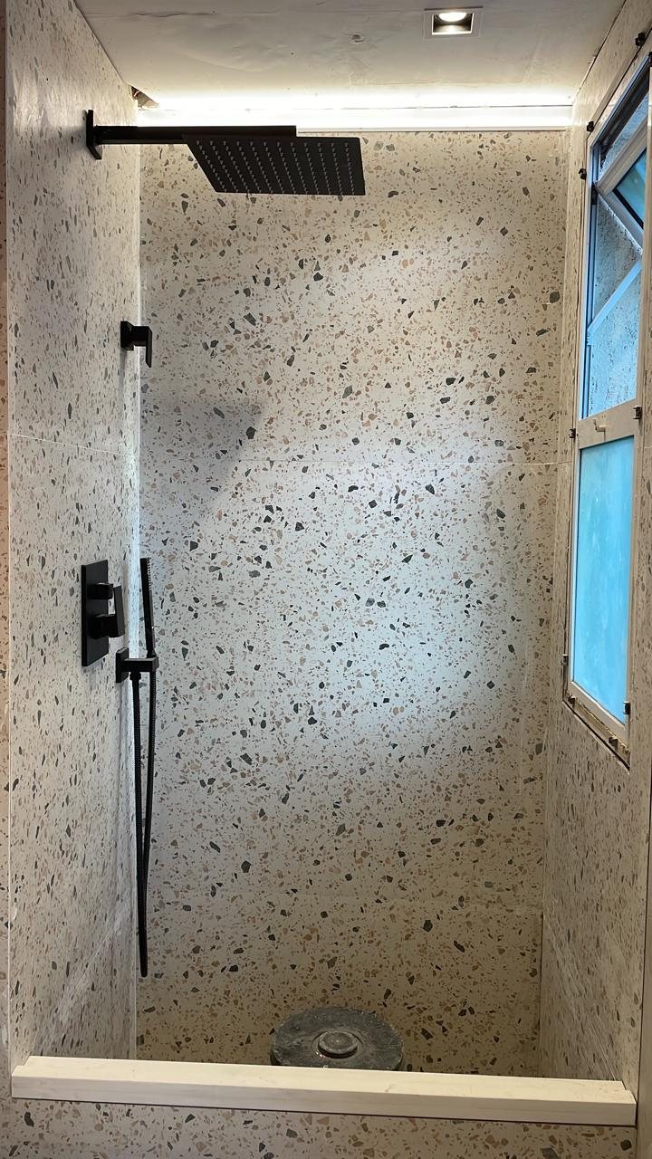 Box de banheiro em terrazzo com janela, chuveiro de teto e metais pretos.