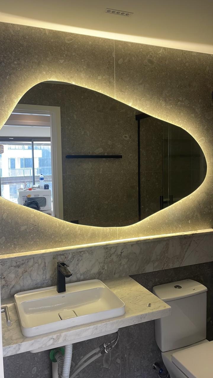Banheiro com espelho orgânico iluminado com LED, bancada de mármore, cuba branca e torneira preta.