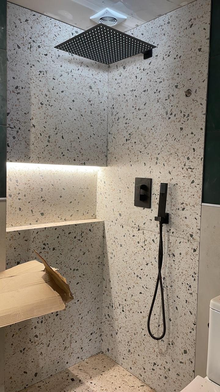 Box de banheiro com revestimento em terrazzo, chuveiro de teto quadrado preto fosco e nicho iluminado.